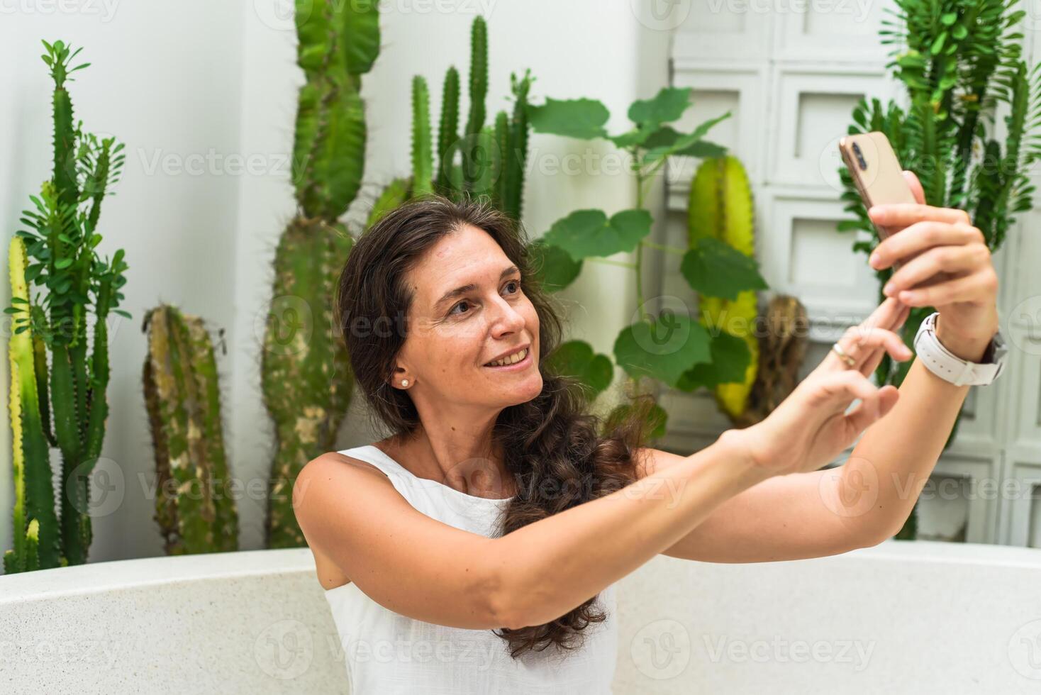 Portrait of 40 years old woman without make up sitting with glass of ice coffee in white dress and taking a selfie photo