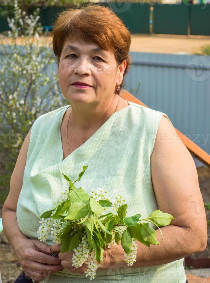 Portrait of 65 years old senior woman in sleeveless blouse with neutral facial expression sitting and holding the spring bouquet of flowering branches of bird cherry tree photo