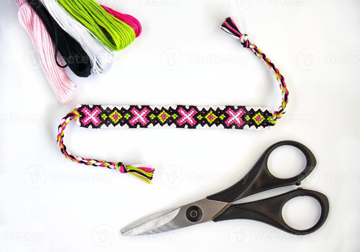 Woven handmade DIY friendship bracelets with bright pink and green pattern next to skeins of thread and scissors on white background photo