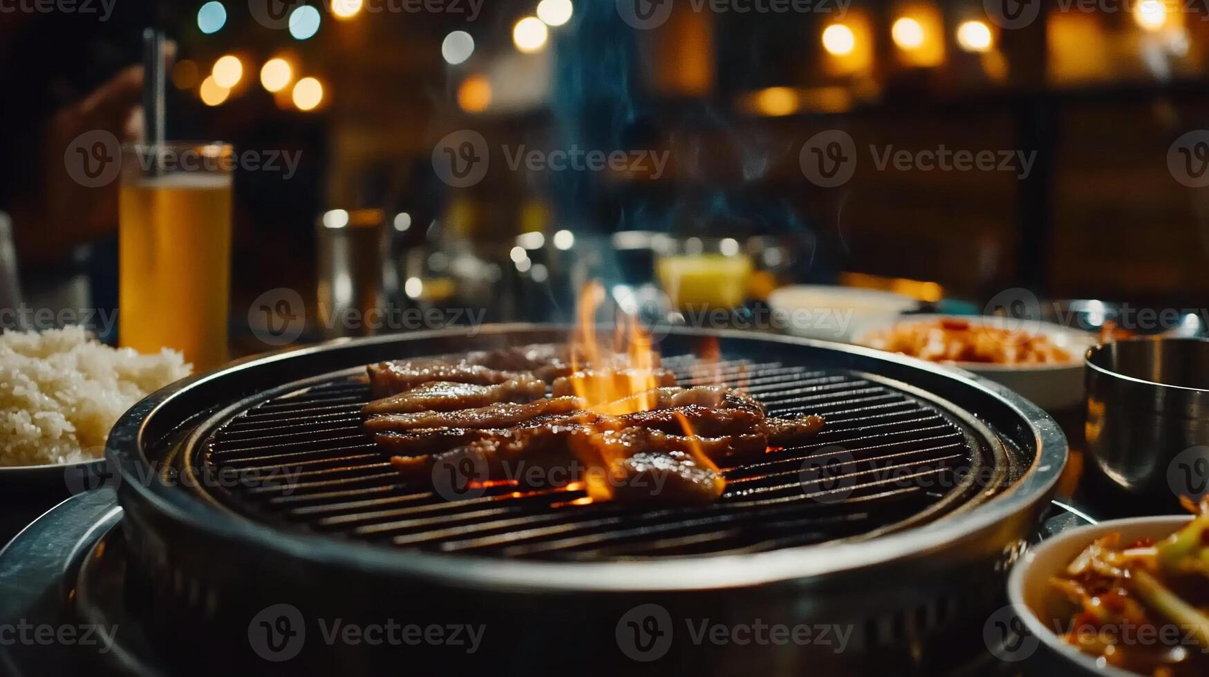 Delicious grilled meat sizzling on a hot grill. Flaring flames and smoke rise from the cooked meat. A variety of side dishes and a glass of beer are visible photo