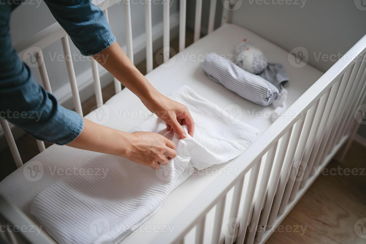 Mother in denim shirt folds white cloth inside a crib, soft toy nearby photo