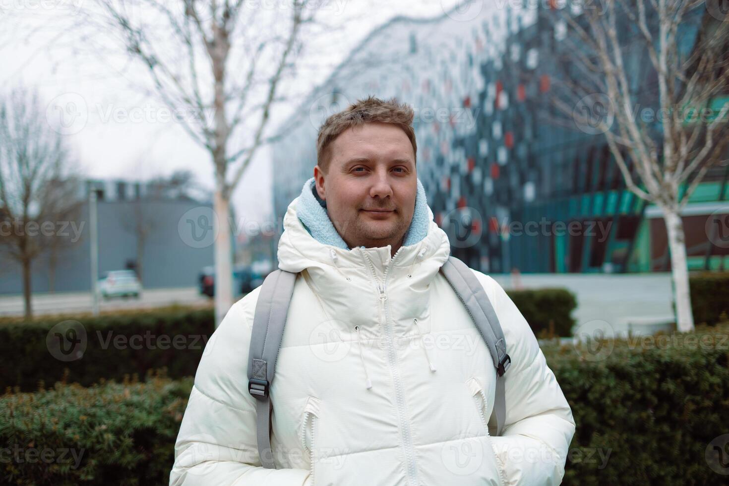 Portrait of Caucasian smiling bearded man with backpack, looking at camera aganist airport terminal building in the winter city. Lifestyle, city life concept photo
