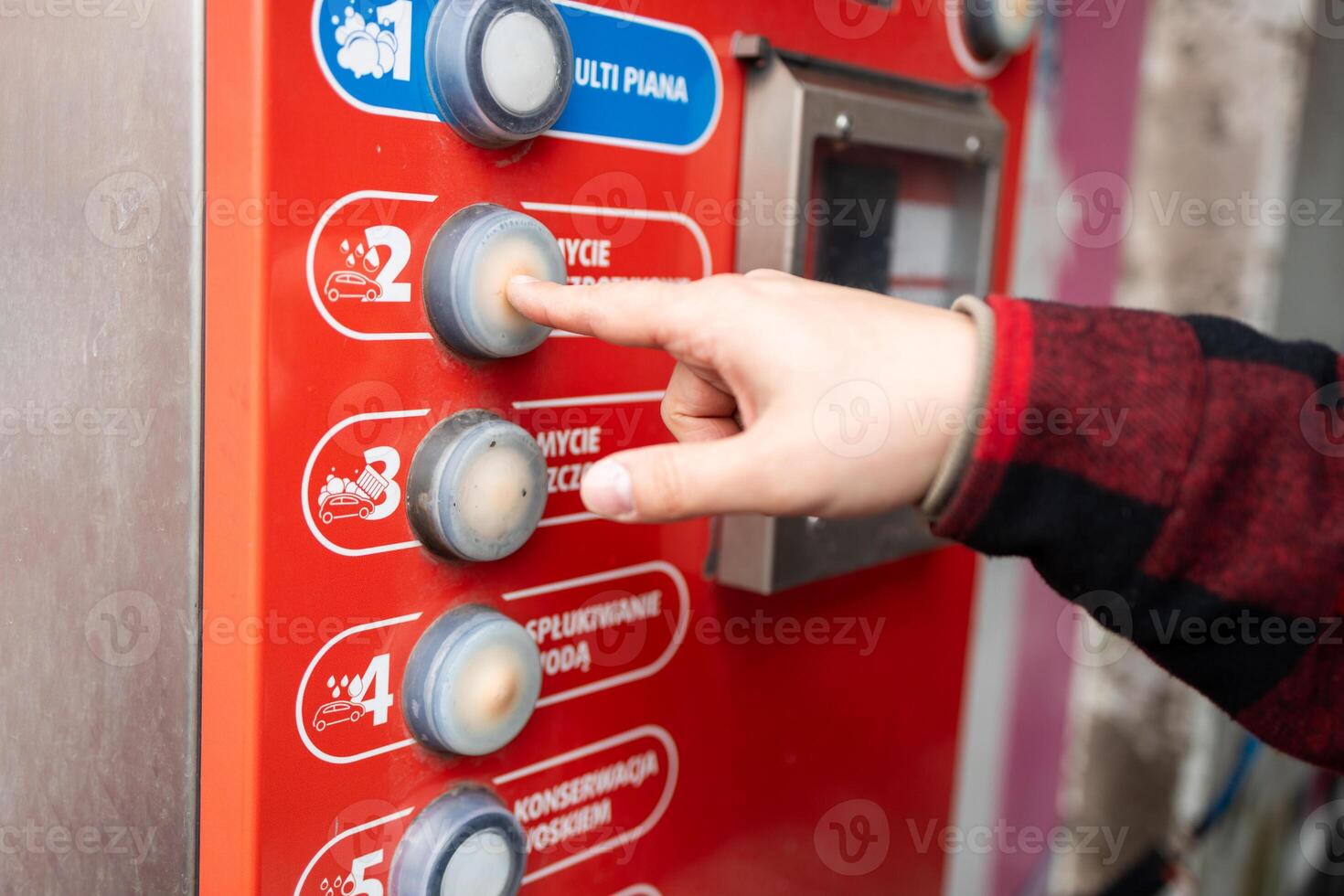 Self service car wash station featuring a red and white theme, a control panel, blue hose, instructional icons, and trees in the background under daylight. photo