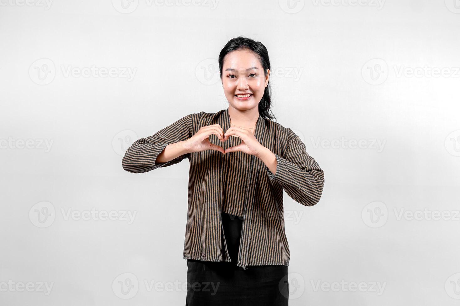 An Indonesian woman wearing traditional lurik attire smiles brightly while forming a heart shape with her hands, showcasing cultural pride and warmth against a neutral background. photo