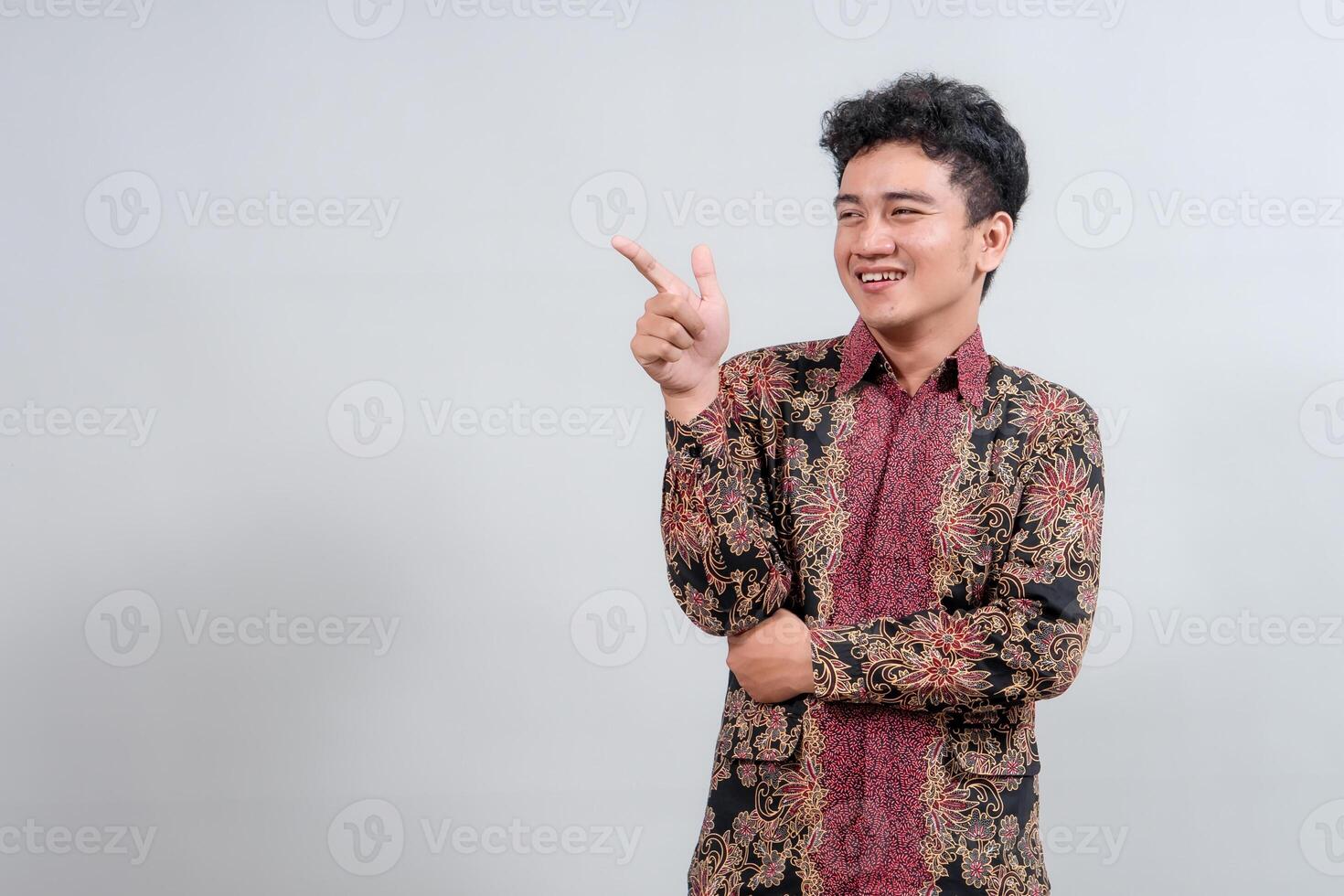 A smiling young man in traditional batik attire points to the side, presenting or highlighting something, ideal for cultural or promotional themes photo