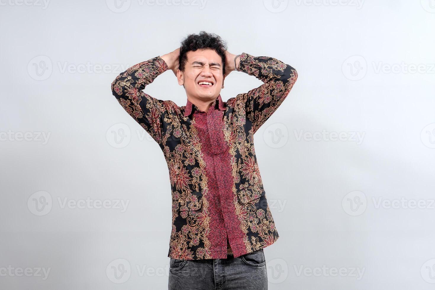 A young man in traditional batik attire scratches his head with a confused expression, depicting forgetfulness or contemplation photo