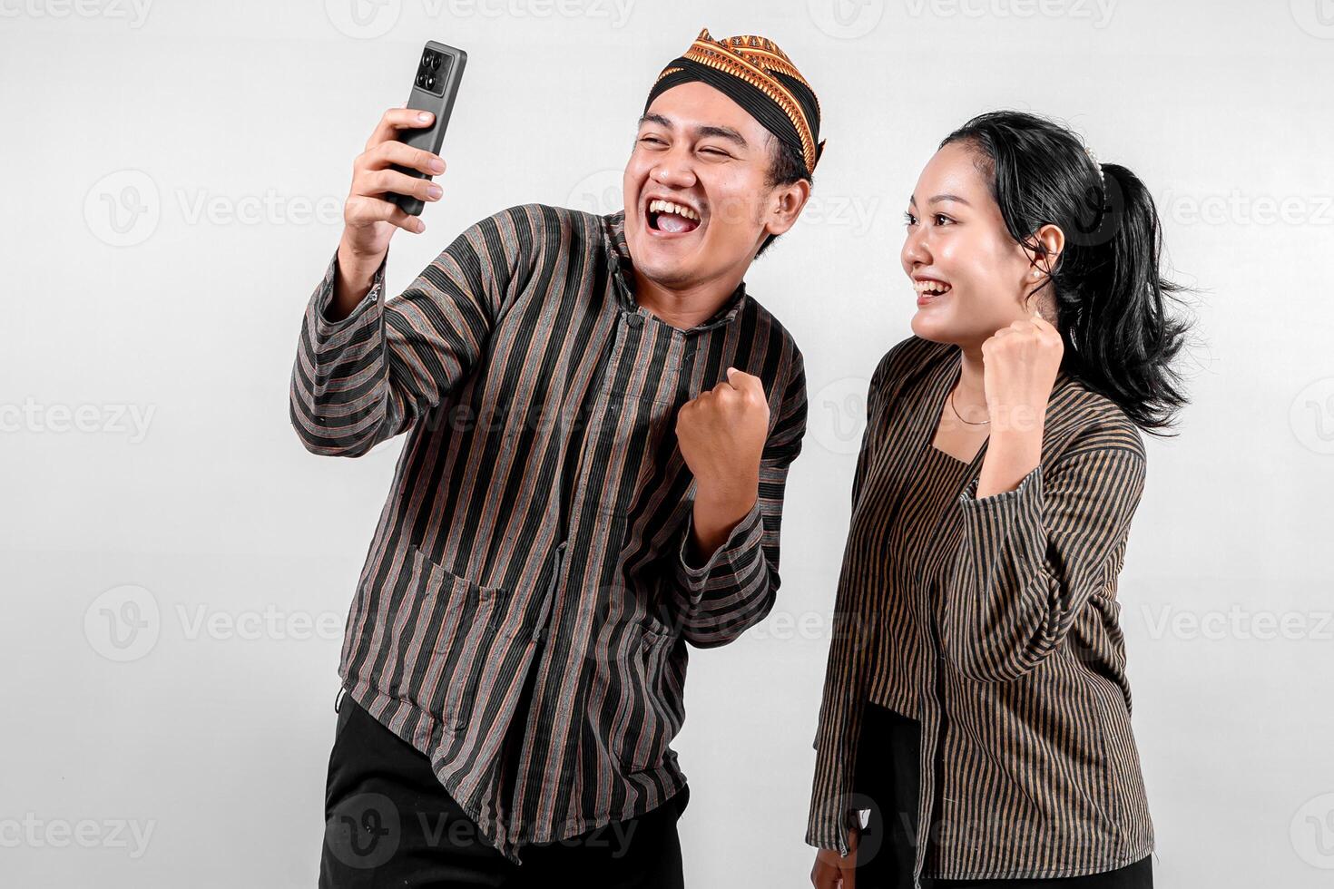 Indonesian couple in traditional Javanese Lurik or Sorjan outfit celebrating success while taking a selfie. Cheerful expression, isolated on white background. photo