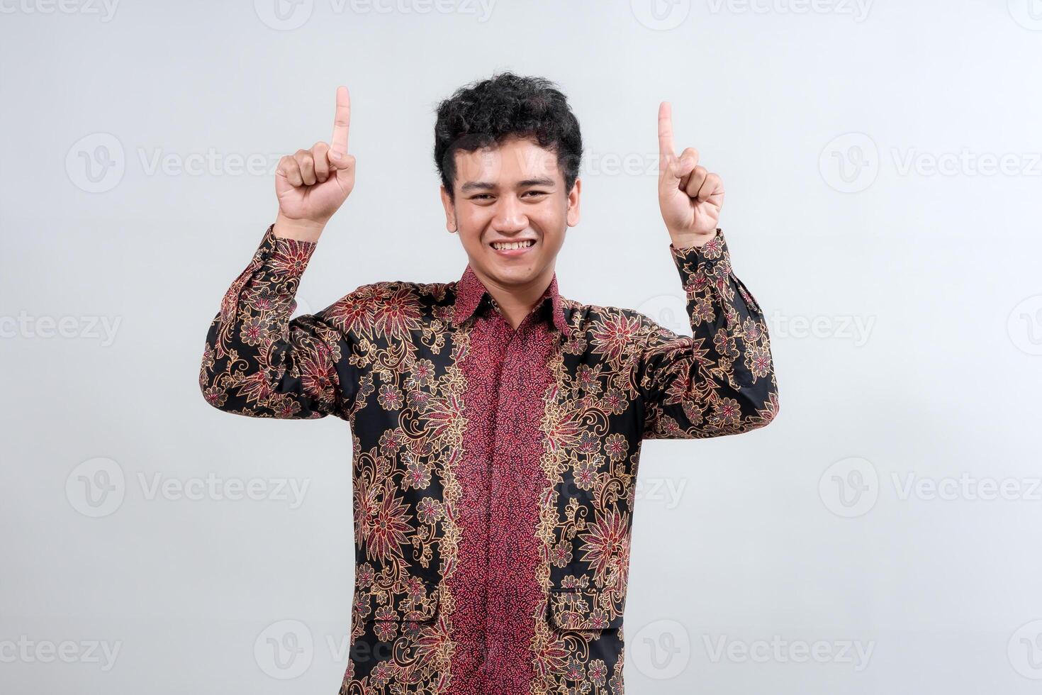 A joyful young man in a traditional batik shirt raises both hands with fingers pointing up, expressing excitement and positivity, perfect for cultural or celebratory themes. photo