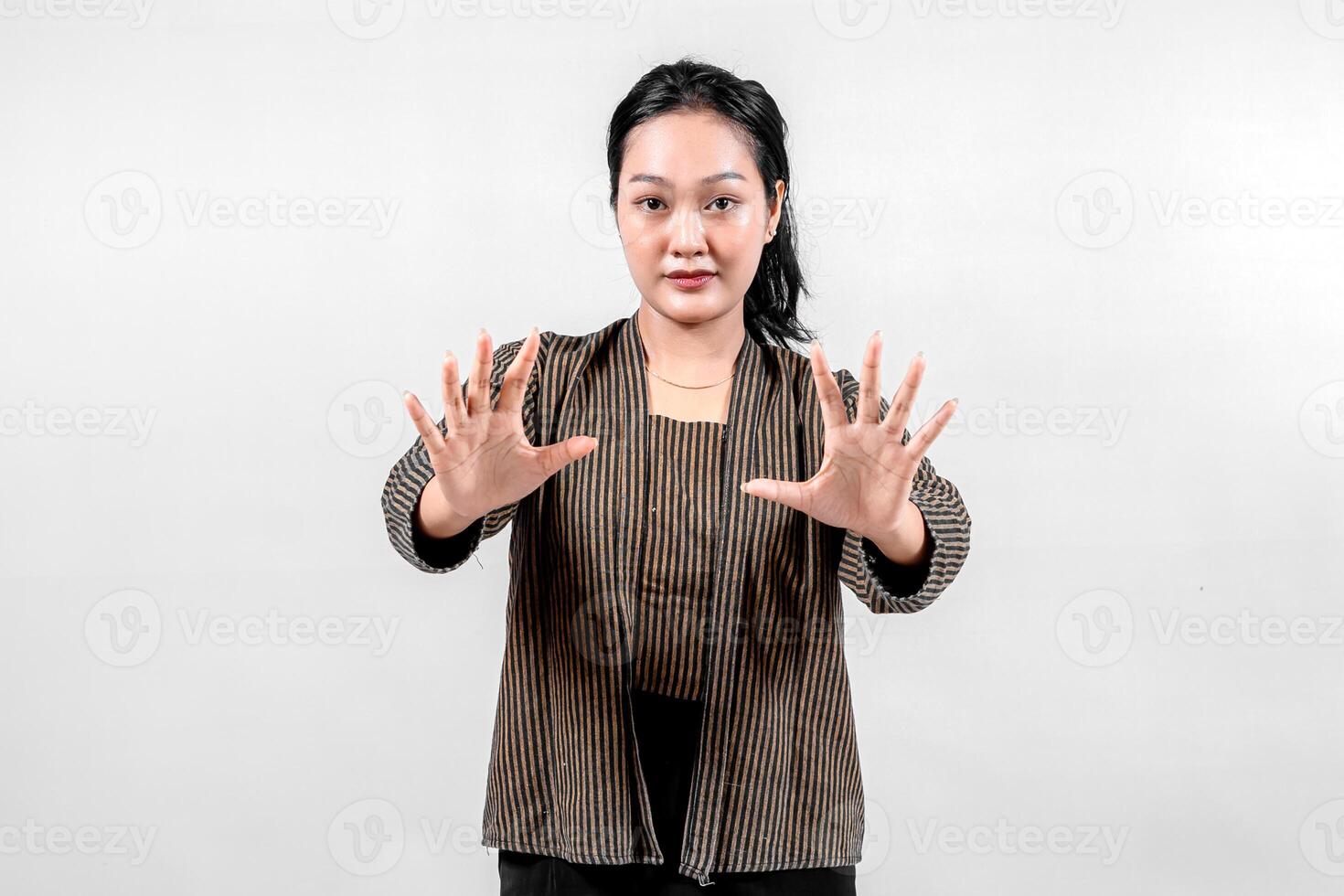 Indonesian woman wearing traditional lurik attire smiles confidently while extending both hands forward with open palms, showcasing cultural pride and a welcoming gesture against a neutral background. photo