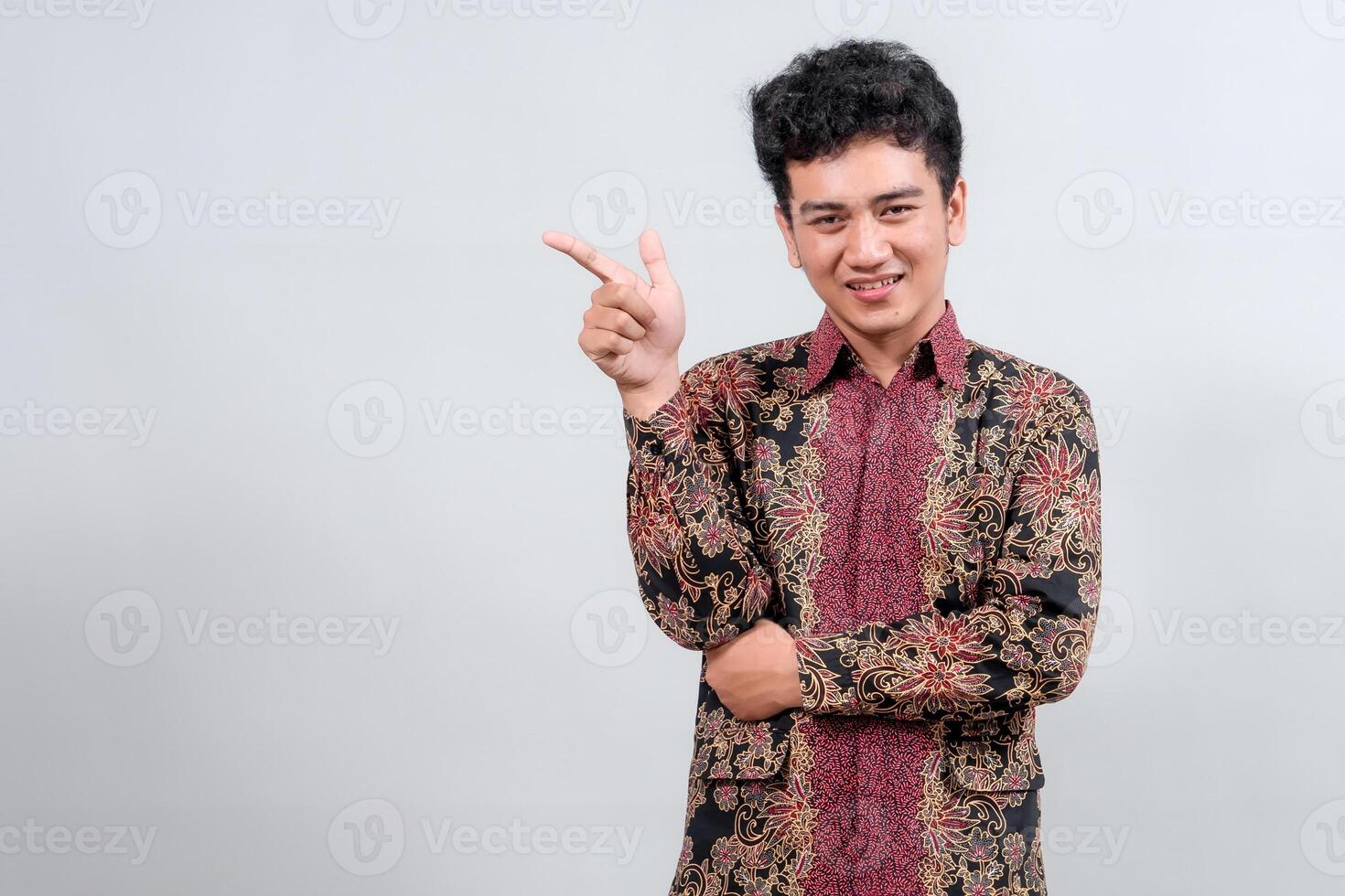 A smiling young man in traditional batik attire points to the side, presenting or highlighting something, ideal for cultural or promotional themes. photo