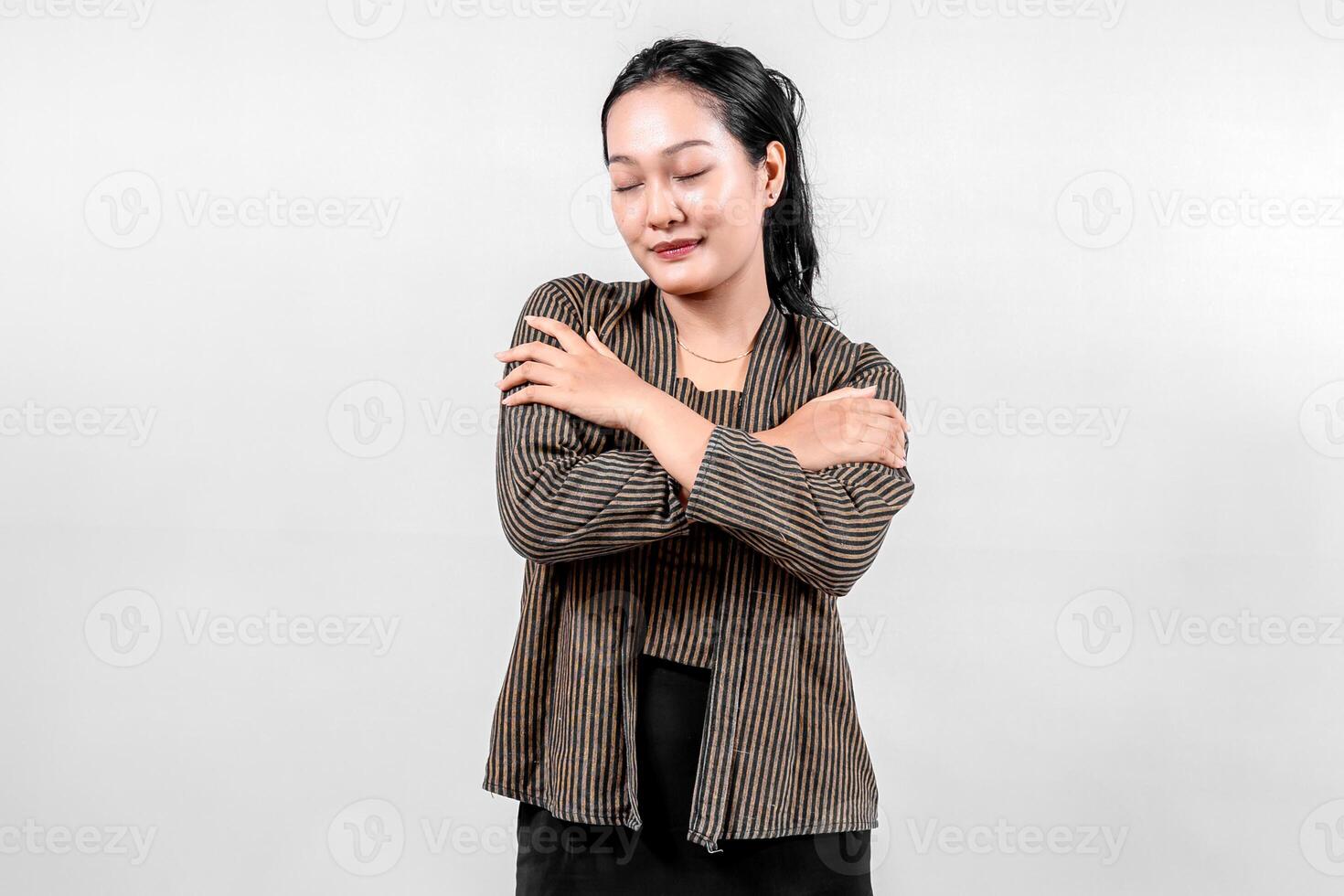 Indonesian woman wearing traditional lurik attire smiles gently with her eyes closed, crossing her arms over her chest, showcasing cultural pride and warmth against a neutral background. photo