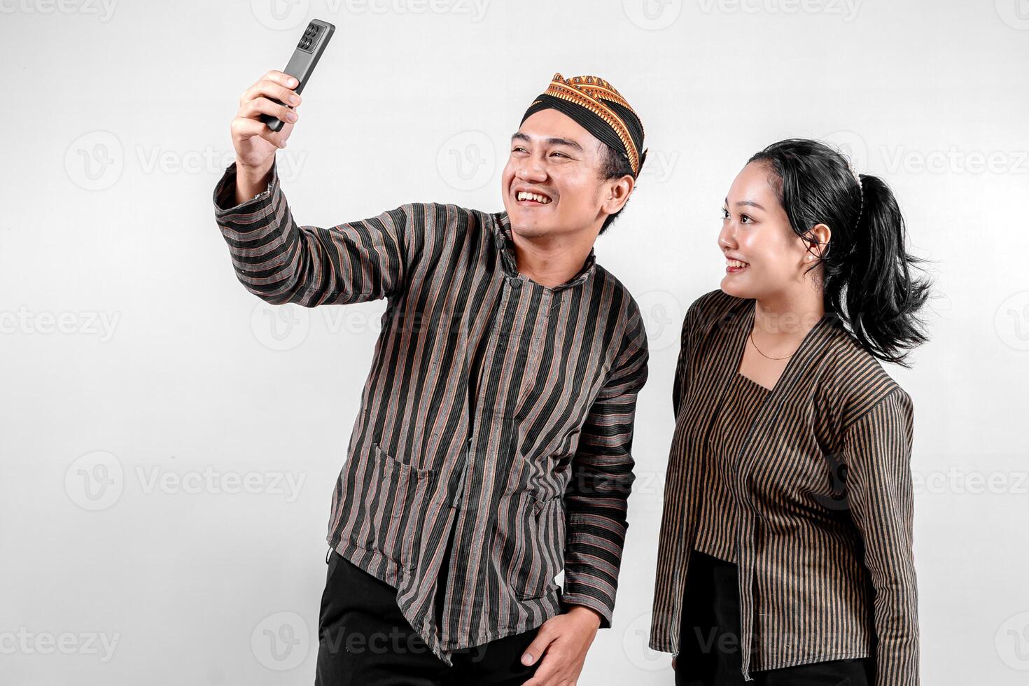 Indonesian couple wearing traditional Javanese Lurik or Sorjan clothing, smiling while taking a selfie. Symbol of heritage, joy, and togetherness. Isolated on white background. photo