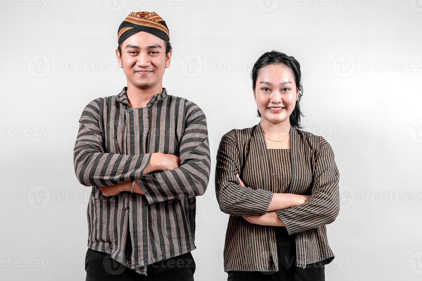 A cheerful Indonesian man and a woman, wearing traditional lurik attire, pose confidently with crossed arms against a plain background, expressing harmony, culture, and confidence. photo
