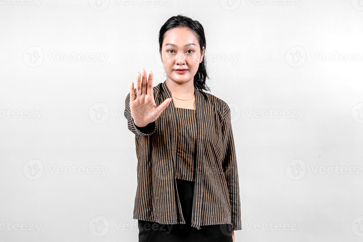 An Indonesian woman wearing traditional lurik attire smiles confidently while holding her hand up in a stop gesture, showcasing cultural pride and assertiveness against a neutral background. photo