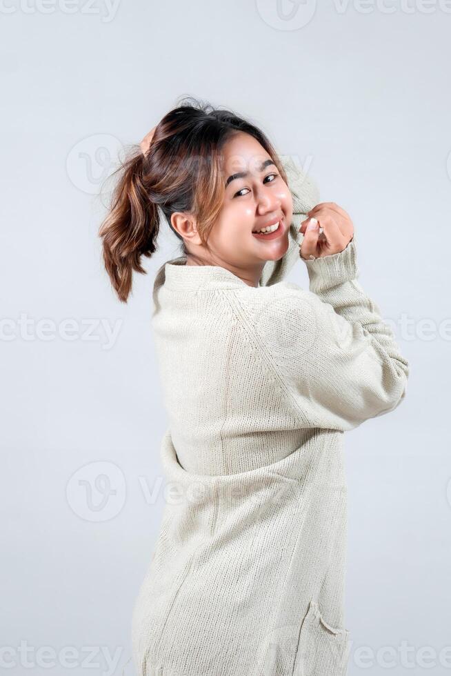 A young woman in a cozy sweater smiles playfully while adjusting her ponytail, radiating a cheerful and casual vibe against a neutral background photo