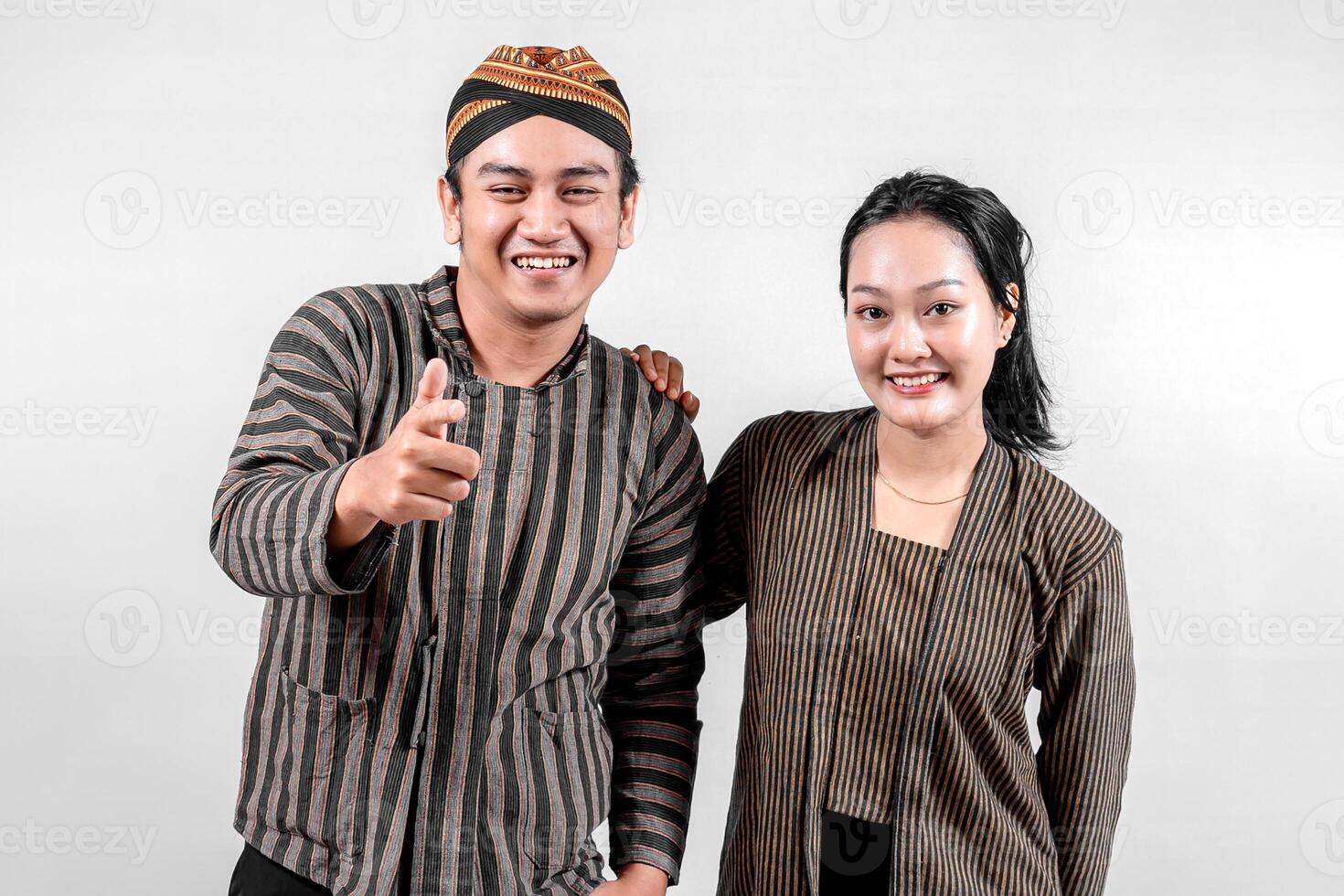 Indonesian couple wearing traditional lurik attire smiles brightly, with the man playfully pointing and the woman standing beside him, showcasing cultural pride and joy against a neutral background photo