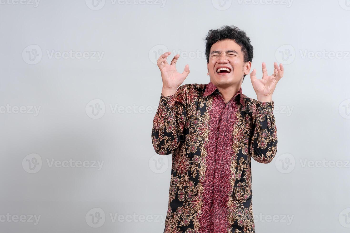 A young man in traditional batik attire expresses playful frustration or excitement with raised hands and a dynamic facial expression, suitable for cultural or emotional themes. photo