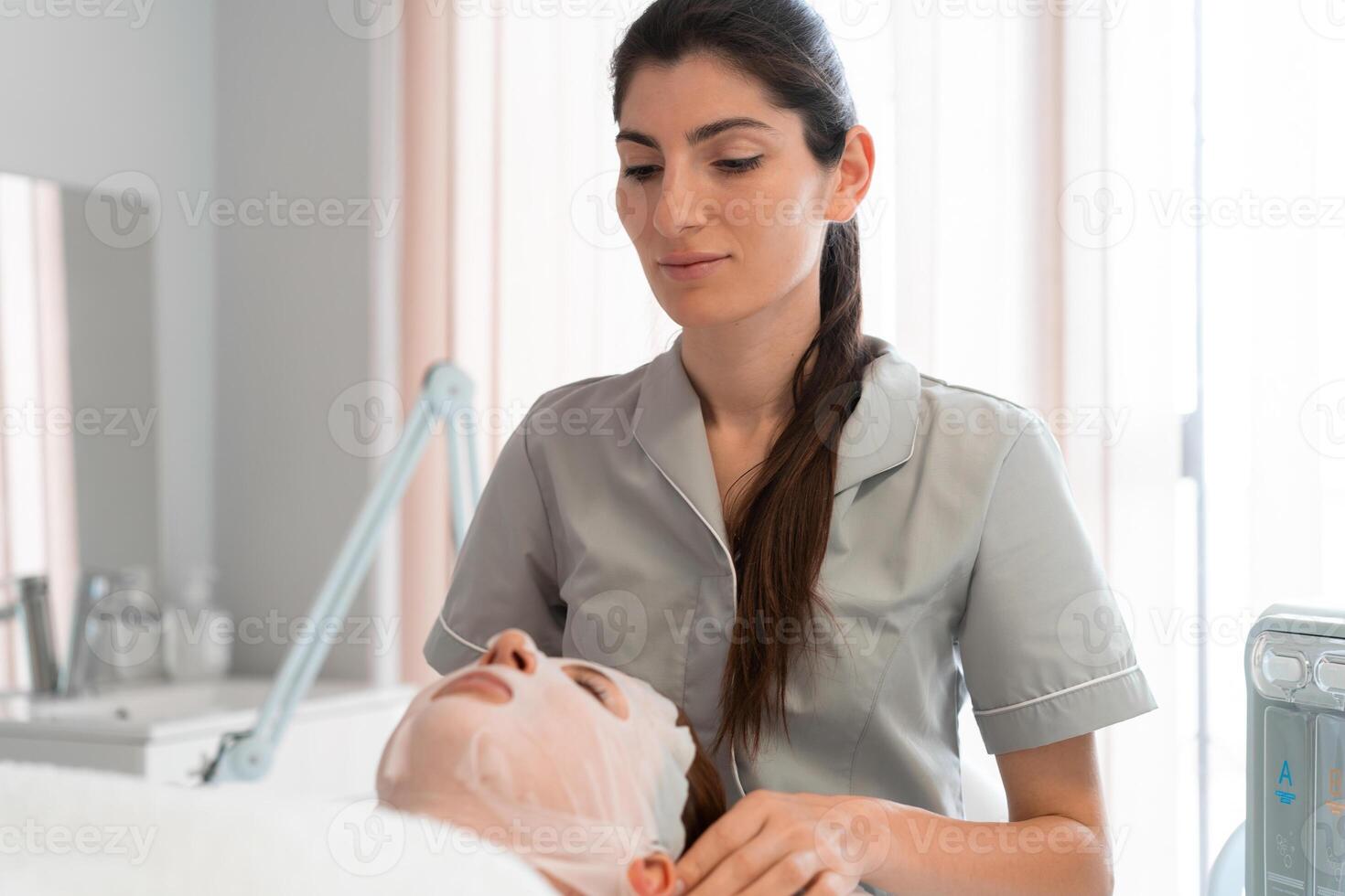 Professional masseuse fixing sheet mask on woman's face at salon photo