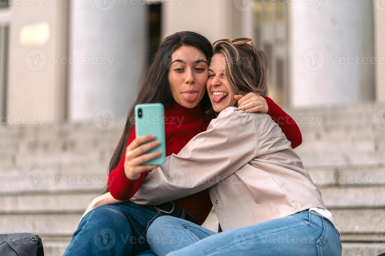 two beautiful student girls taking a selfie sticking out their tongues sitting on the stairs of the college. photo
