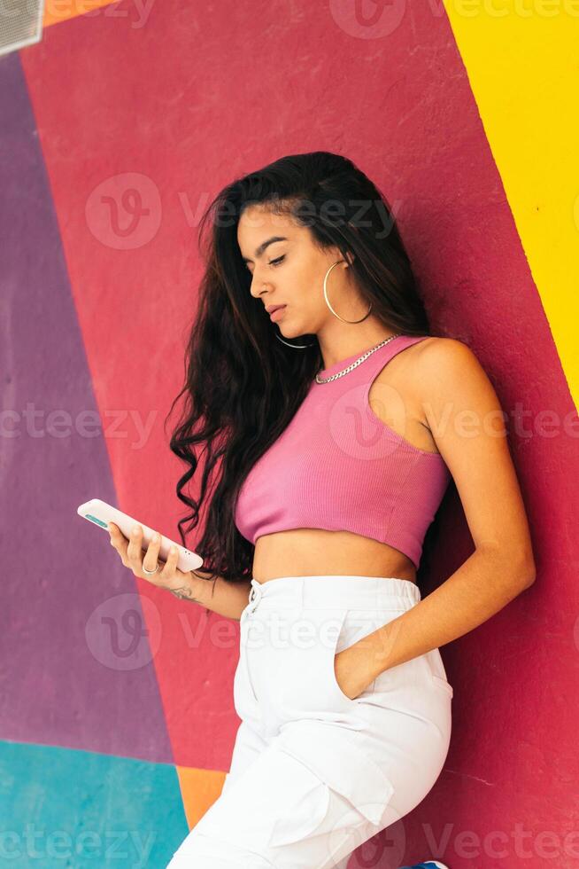 Latin woman using smartphone leaning on a colorful wall photo