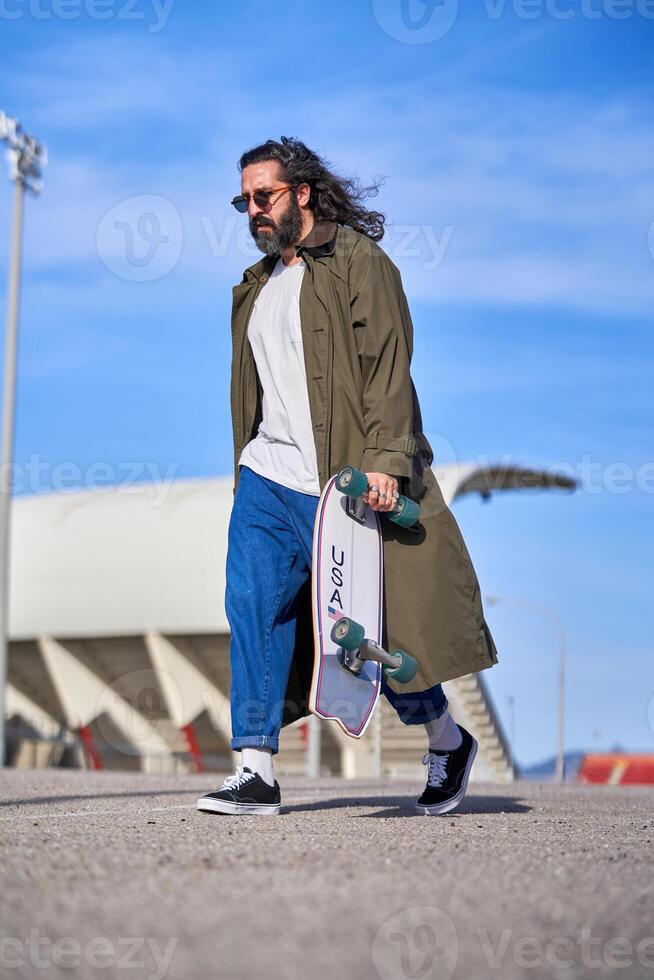 Stylish adult man in raincoat walking on street with skateboard photo