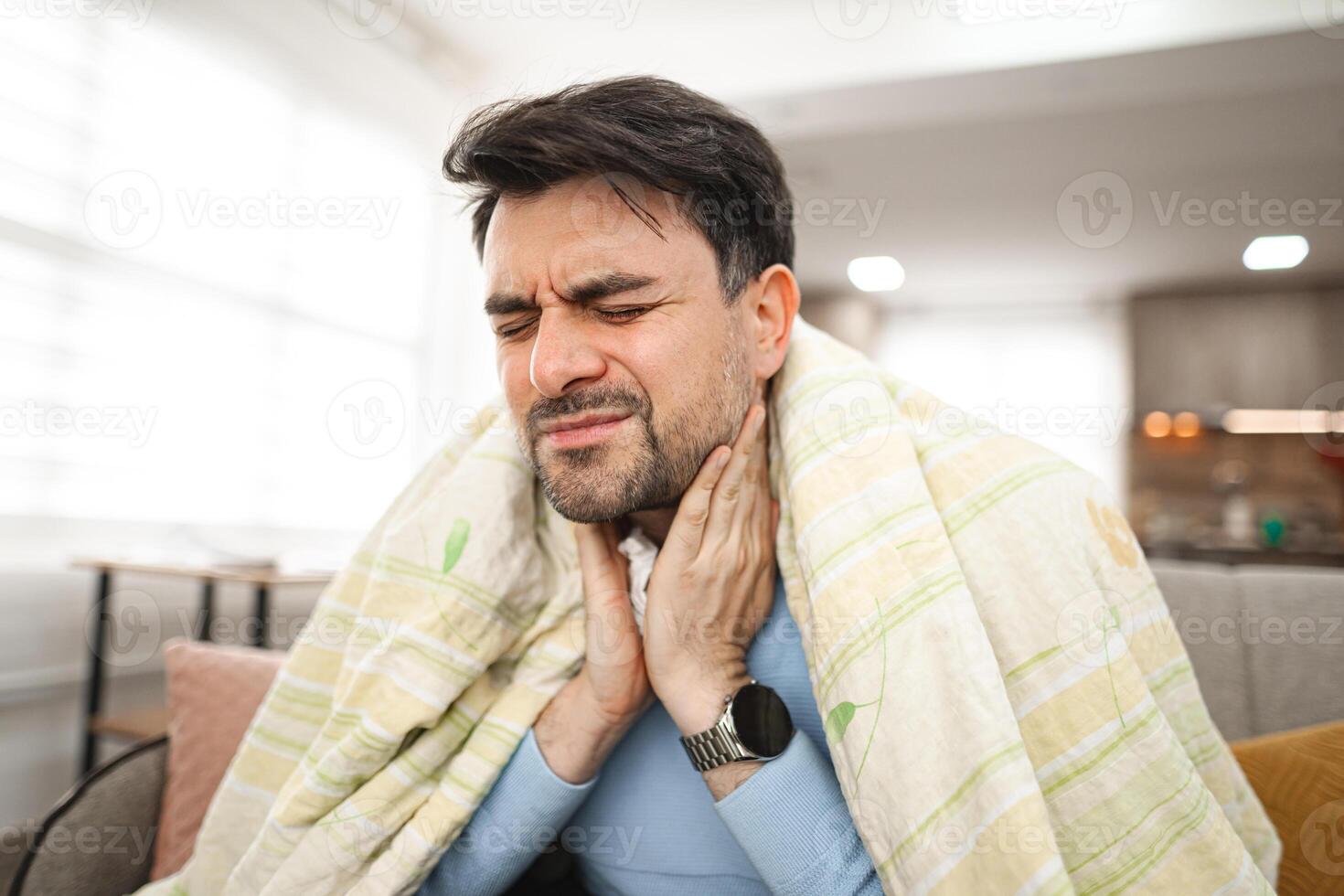 Sick man covered with a blanket is sitting and holding his sick throat. photo