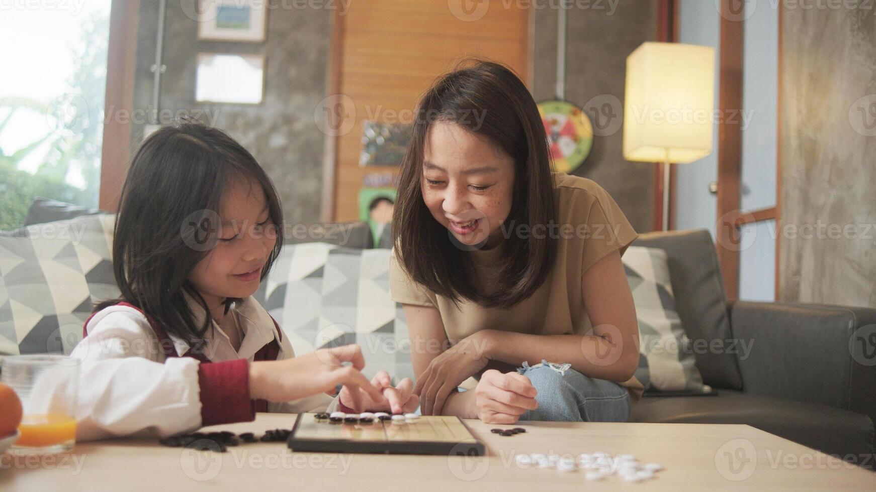 Asian family, mother, and her girl fun playing Go, checkers board game on table in home living room together with love and cheerful, happy weekend relaxing hobby of kid and parent. photo