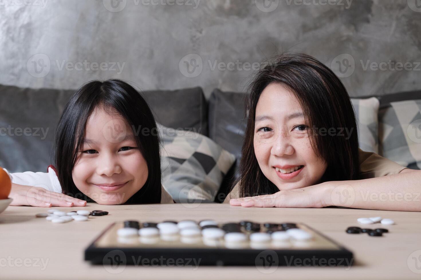 Portrait of Asian mum, and kid fun playing Go, checkers board game on table in home living room with love and cheerful, happy weekend family times relaxing hobby, smile and look at camera together. photo