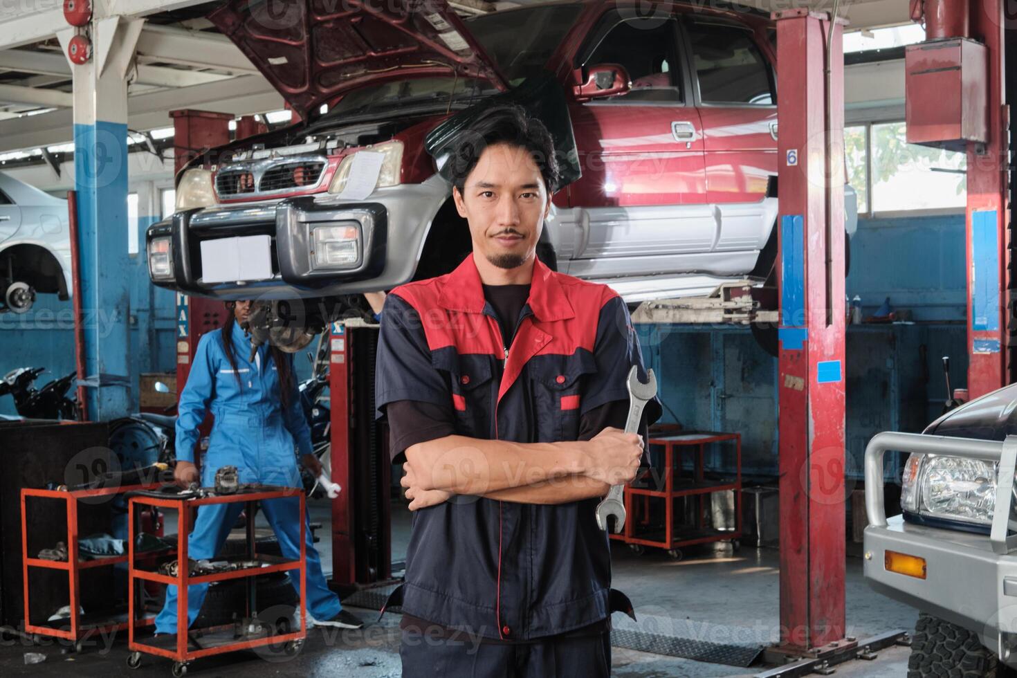 Portrait of Asian male professional mechanic with wrench, arms crossed stands in car fix garage, serious maintenance service jobs, and machine engineer work in automotive transport industry station. photo