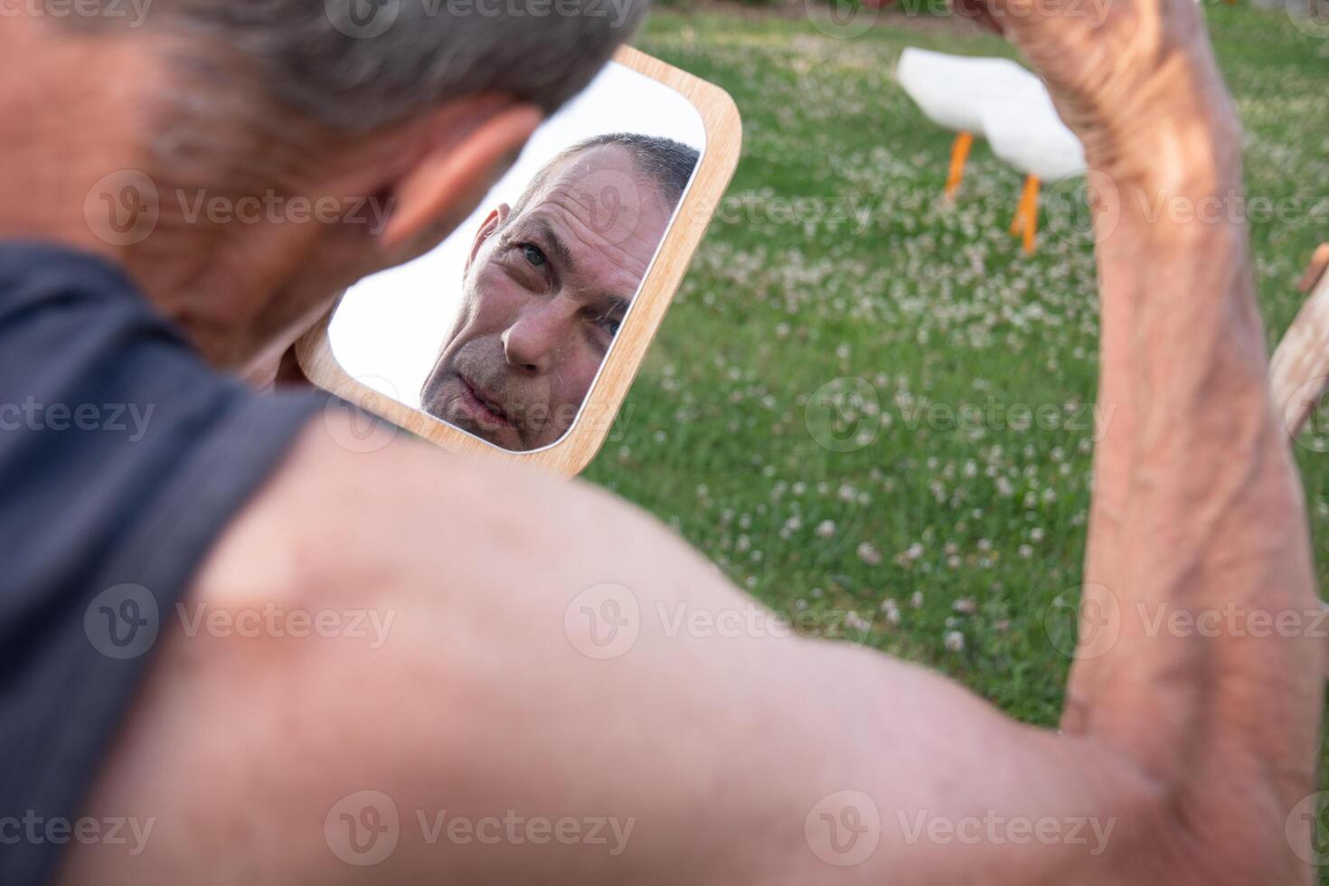 Gray-haired man shaving his hair with clippers in the garden on a green lawn and looking at the mirror, self-care and personal hygiene concept, photo