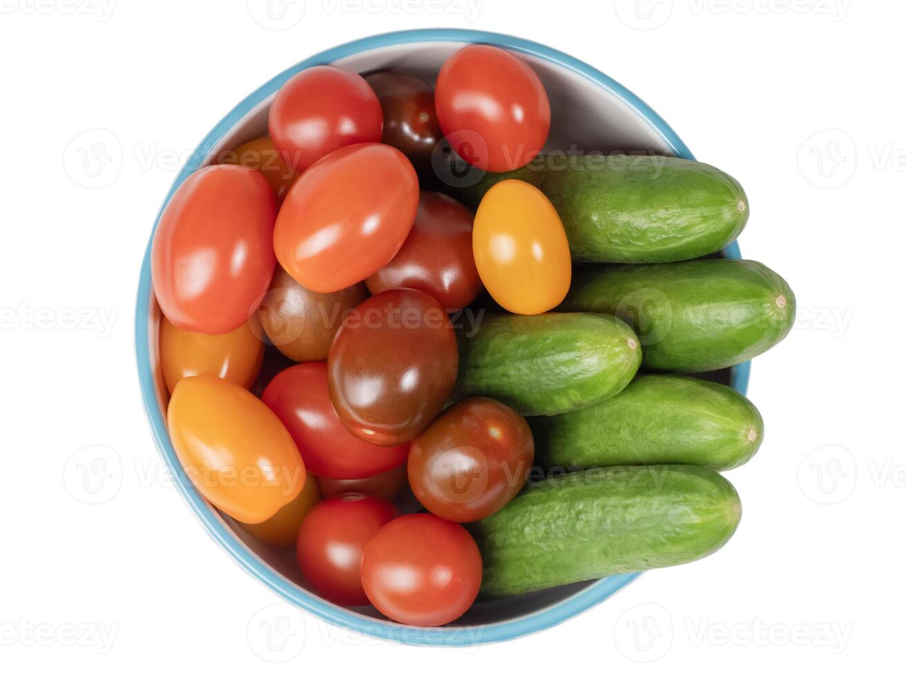 Small colorful cherry tomatoes and fresh green cucumbers overflowing in bowl with blue border, top view, abundant harvest concept, isolated on white background, photo
