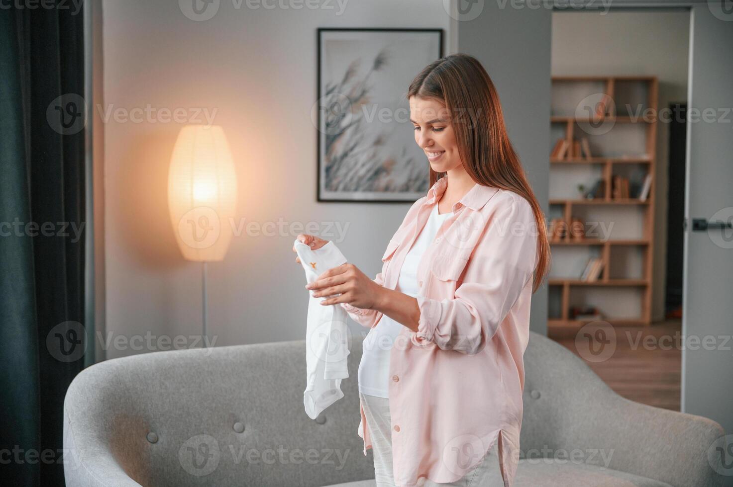 Standing, smiling and holding little shirt. Beautiful pregnant woman is indoors at home photo