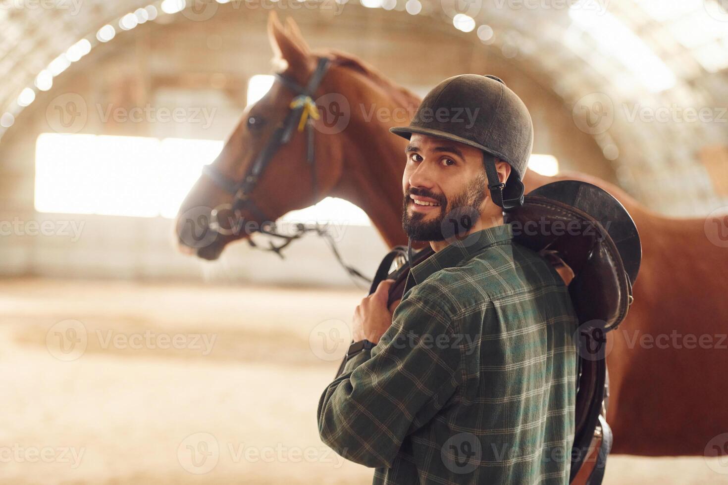 In protective hat. Jockey equipment. Young man with a horse is in the hangar photo