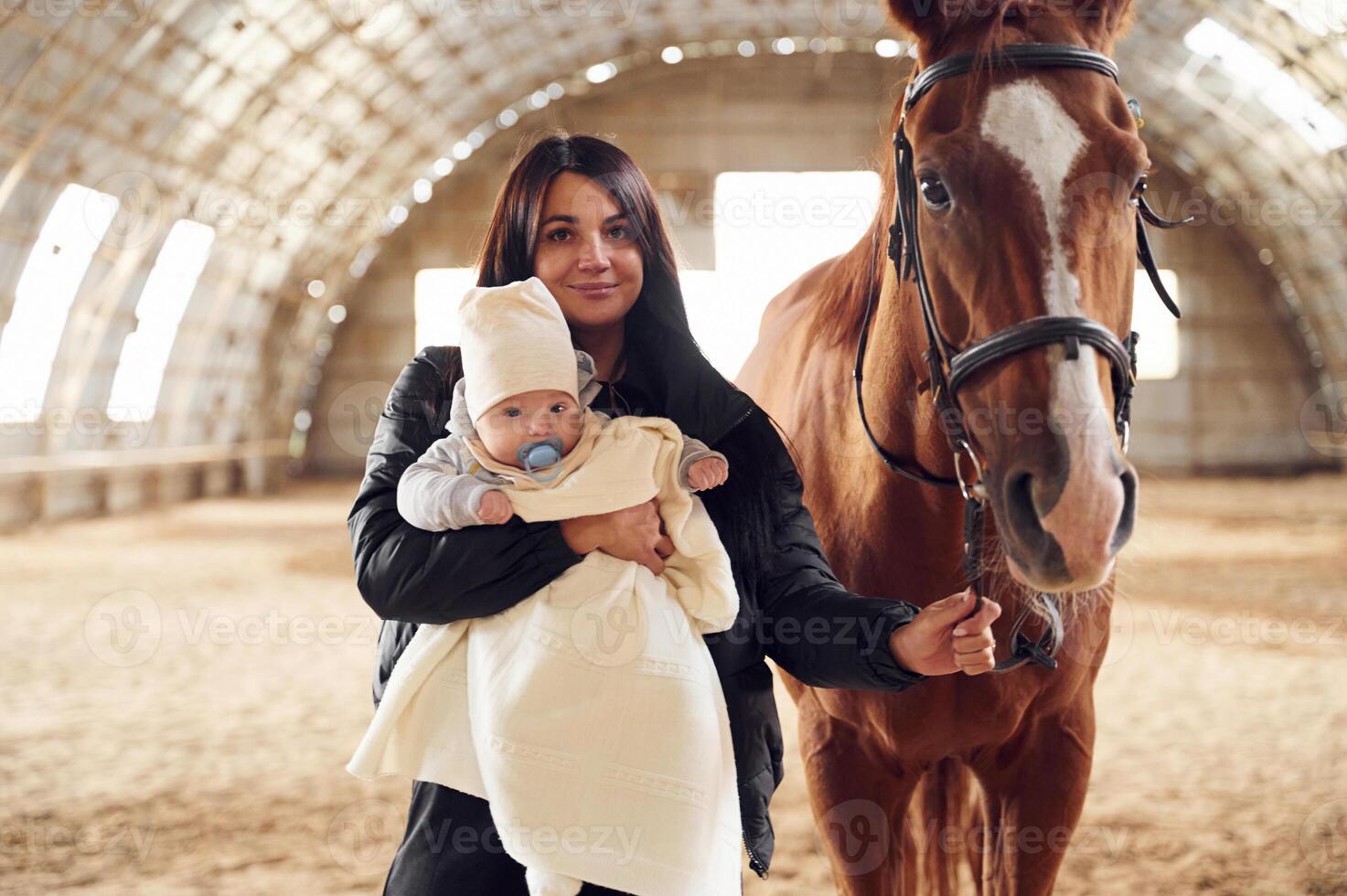 Mother with, baby on the hands is standing indoors with horse in hangar photo