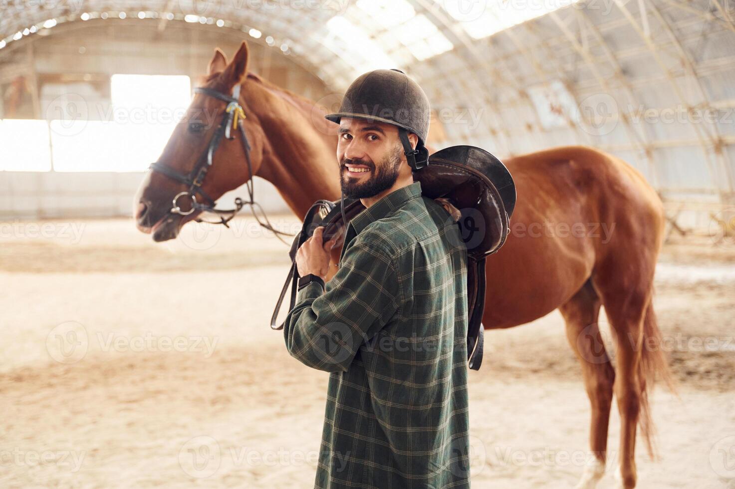 Holding the ,saddle in hands.. Young man with a horse is in the hangar photo