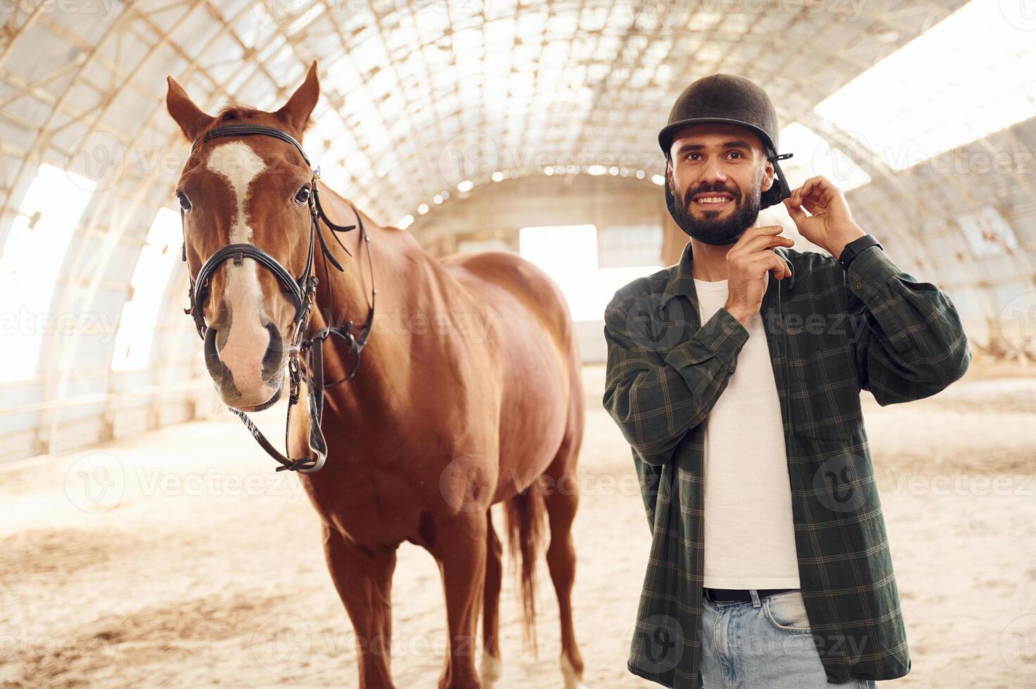 Wearing protective hat and smiling. Young man ,with a horse is in the hangar photo