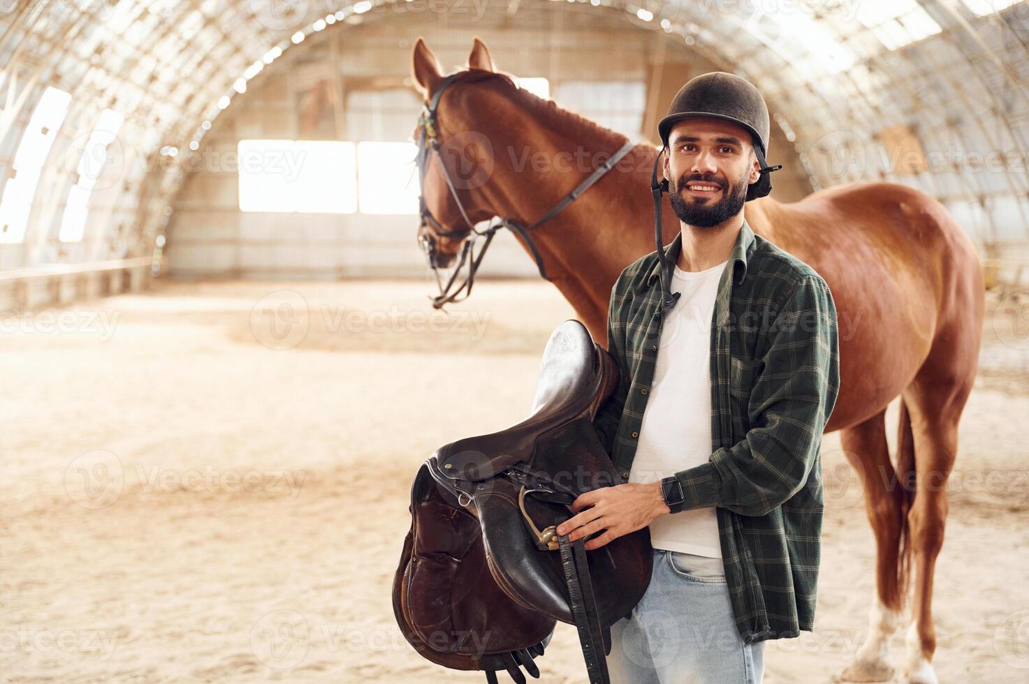 Holding ,the saddle in hands. Young ,man with a horse is in the hangar photo