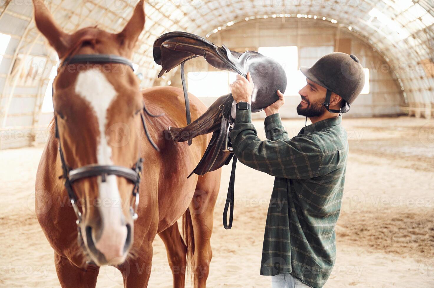 With saddle in hands. Young man with a horse is in the hangar photo