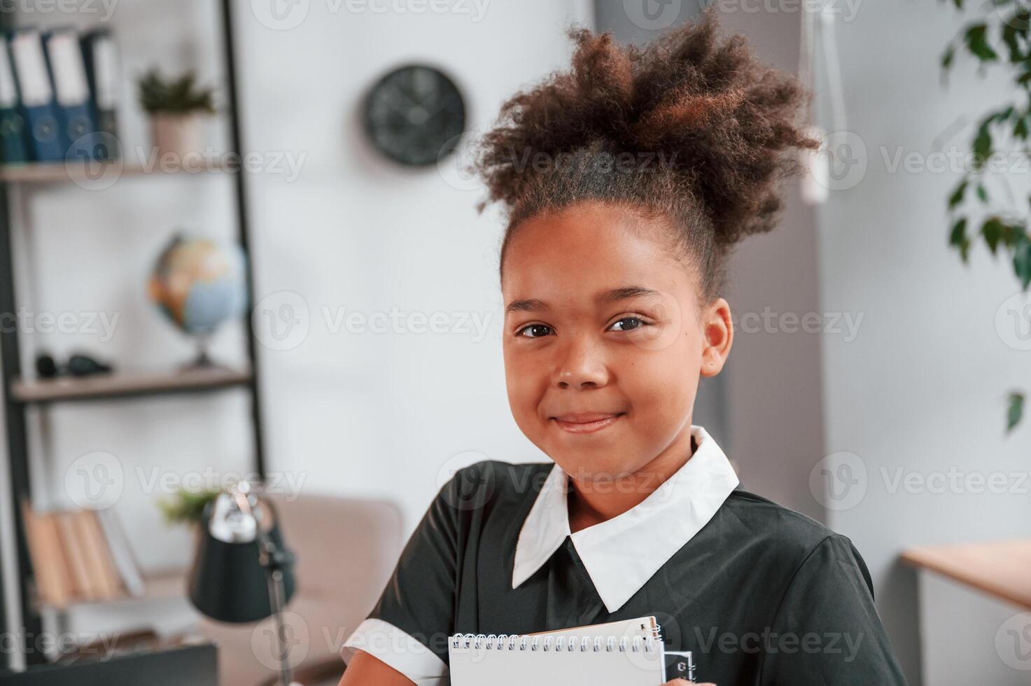 Holding notepad and smiling. Cute little girl in school uniform is standing indoors in domestic room photo