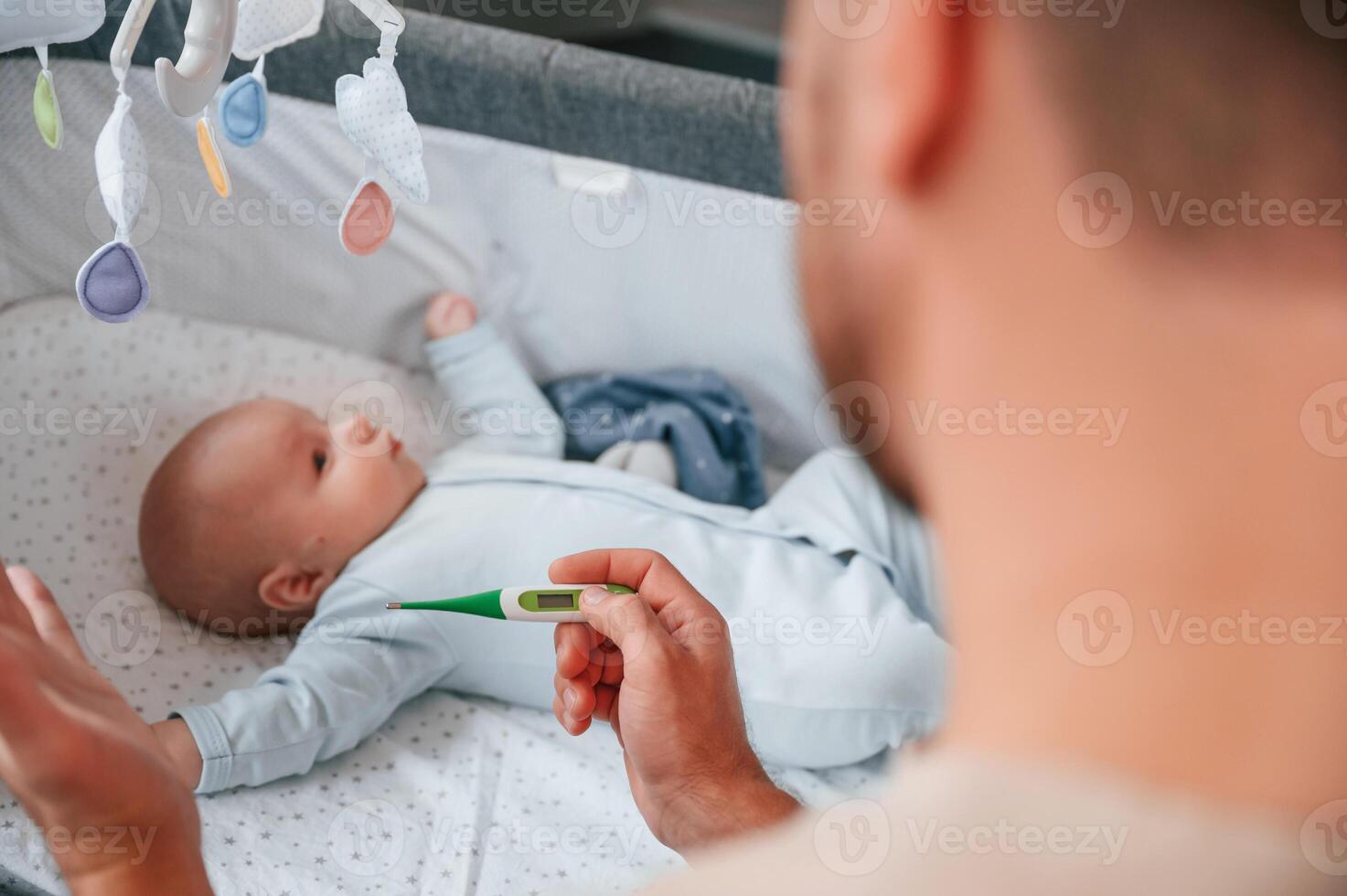 Holding thermometer, kid in the cradle. Father with his newborn baby is indoors. Conception of single dad. photo