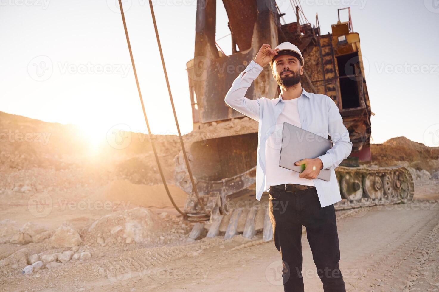 Standing ,against big excavator and holding laptop. Man in uniform is working in the quarry at daytime photo