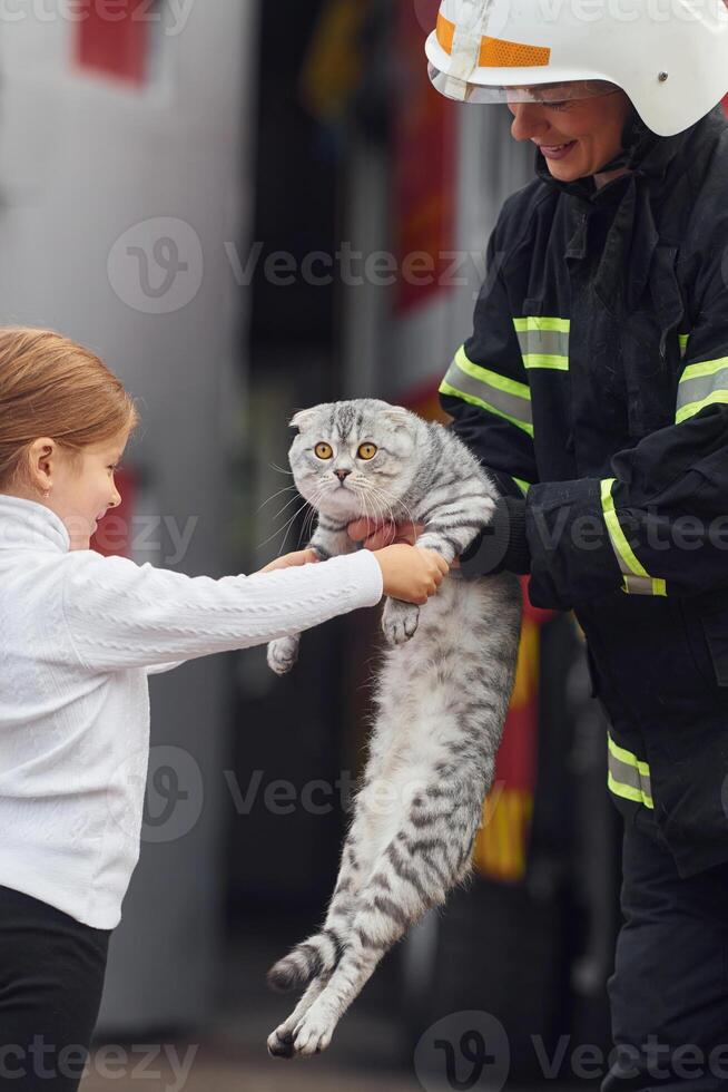 Giving scottish, fold cat to the owner. Firefighter woman in uniform is with a little girl photo