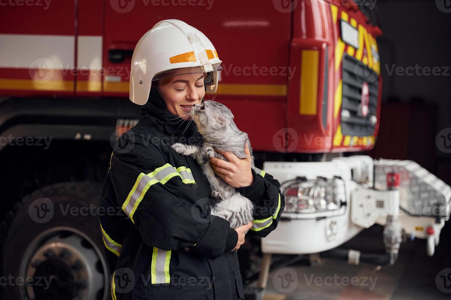 Standing against big truck ,and holding beautiful cat. Woman firefighter in uniform is at work in department photo