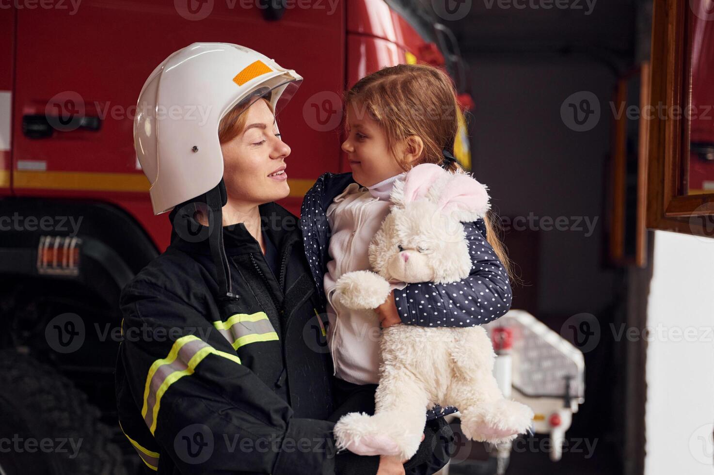 Kid is holding bunny toy. Firefighter woman ,in uniform is with a little girl photo