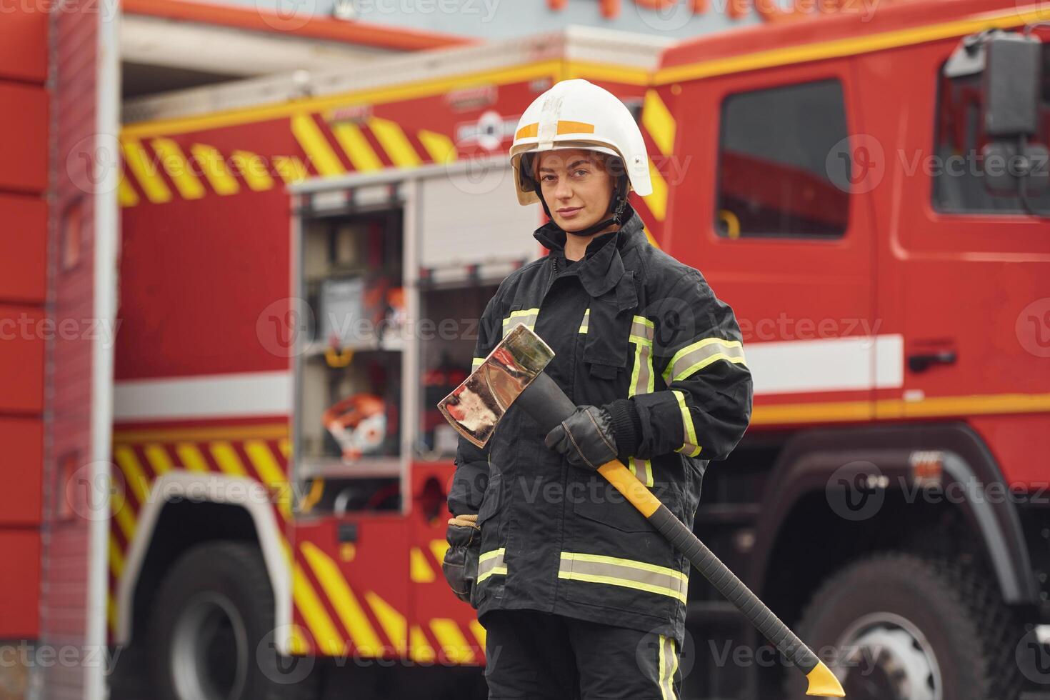 Standing, with axe in hands against truck. Woman firefighter in uniform is at work in department photo