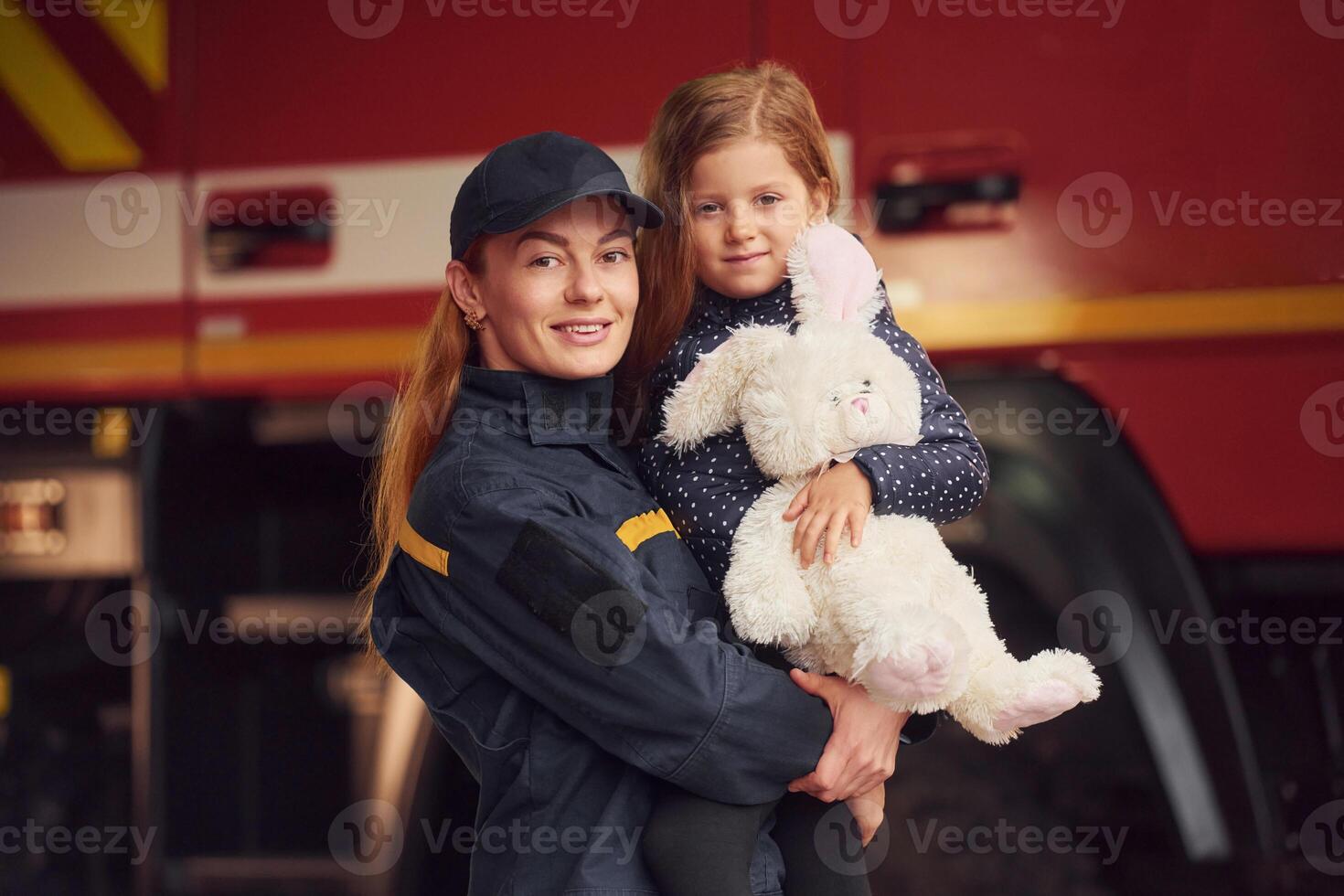 Against big, truck. With bunny toy. Firefighter woman in uniform is with a little girl photo