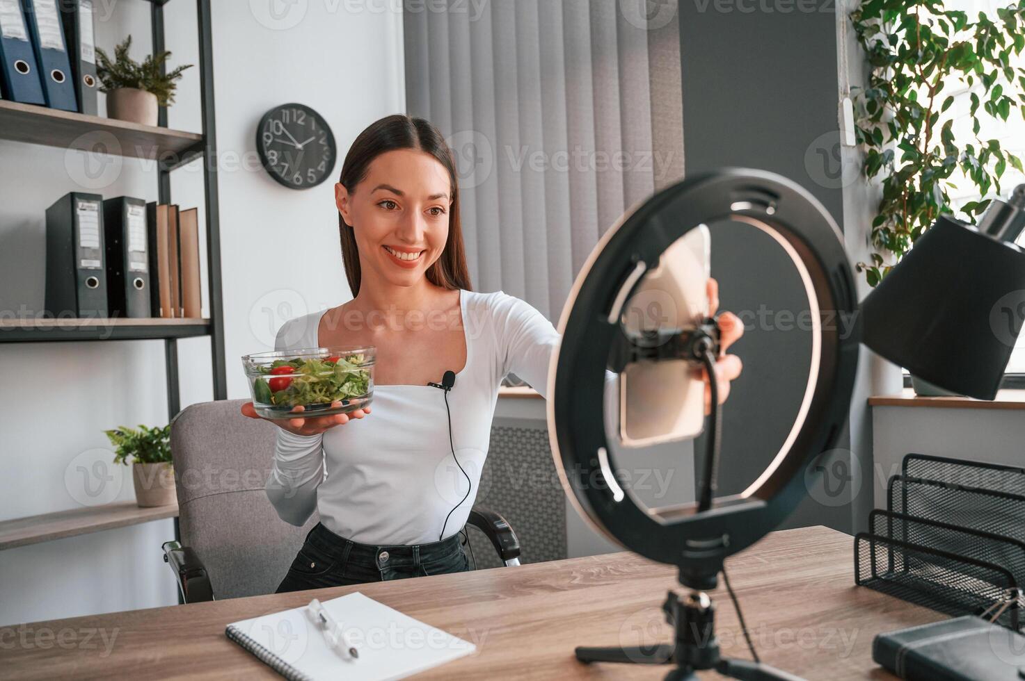 Nutritionist, is showing ,salad on the camera. Young beautiful woman in white clothes is in the office photo