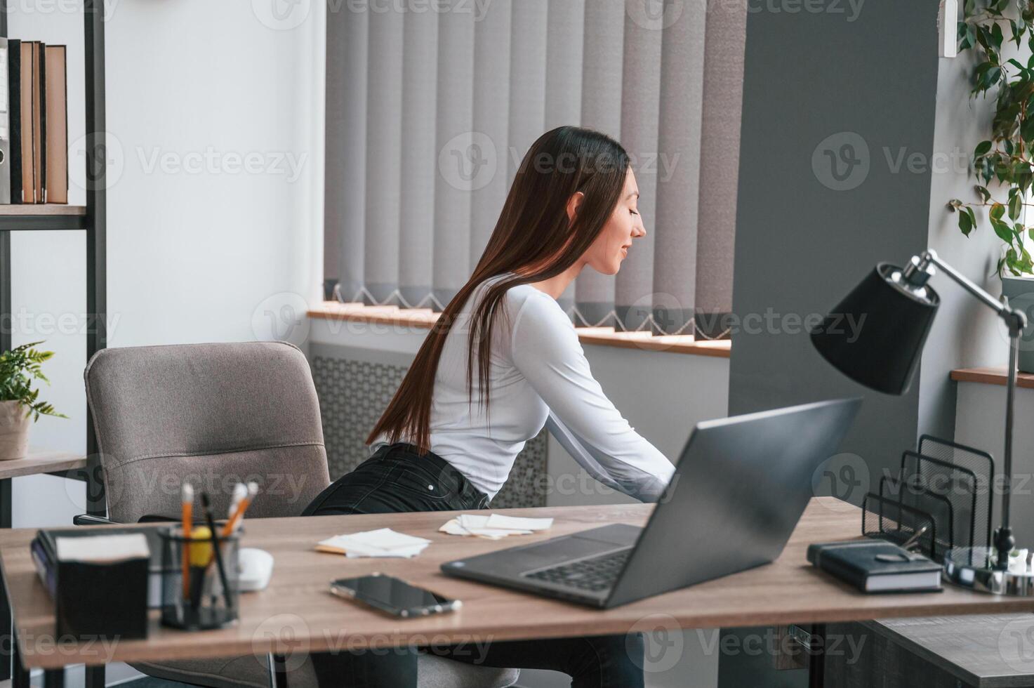 Young beautiful woman in white clothes is in the office and sitting by the table with laptop photo
