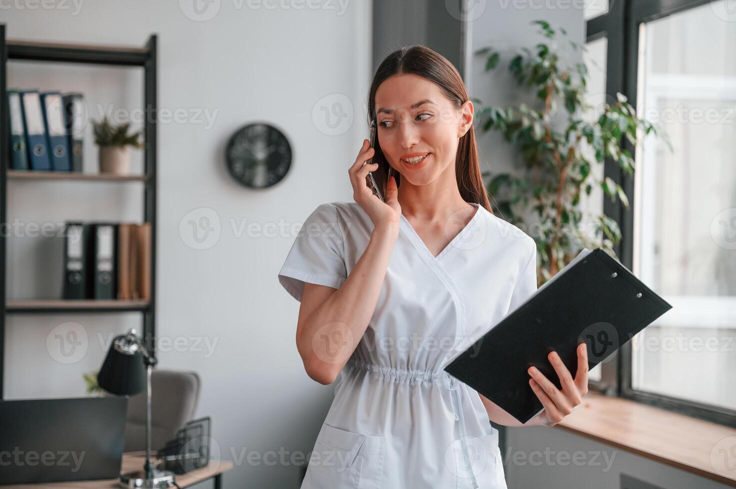 Standing, talking, by phone and holding notepad. Young beautiful woman in white clothes photo