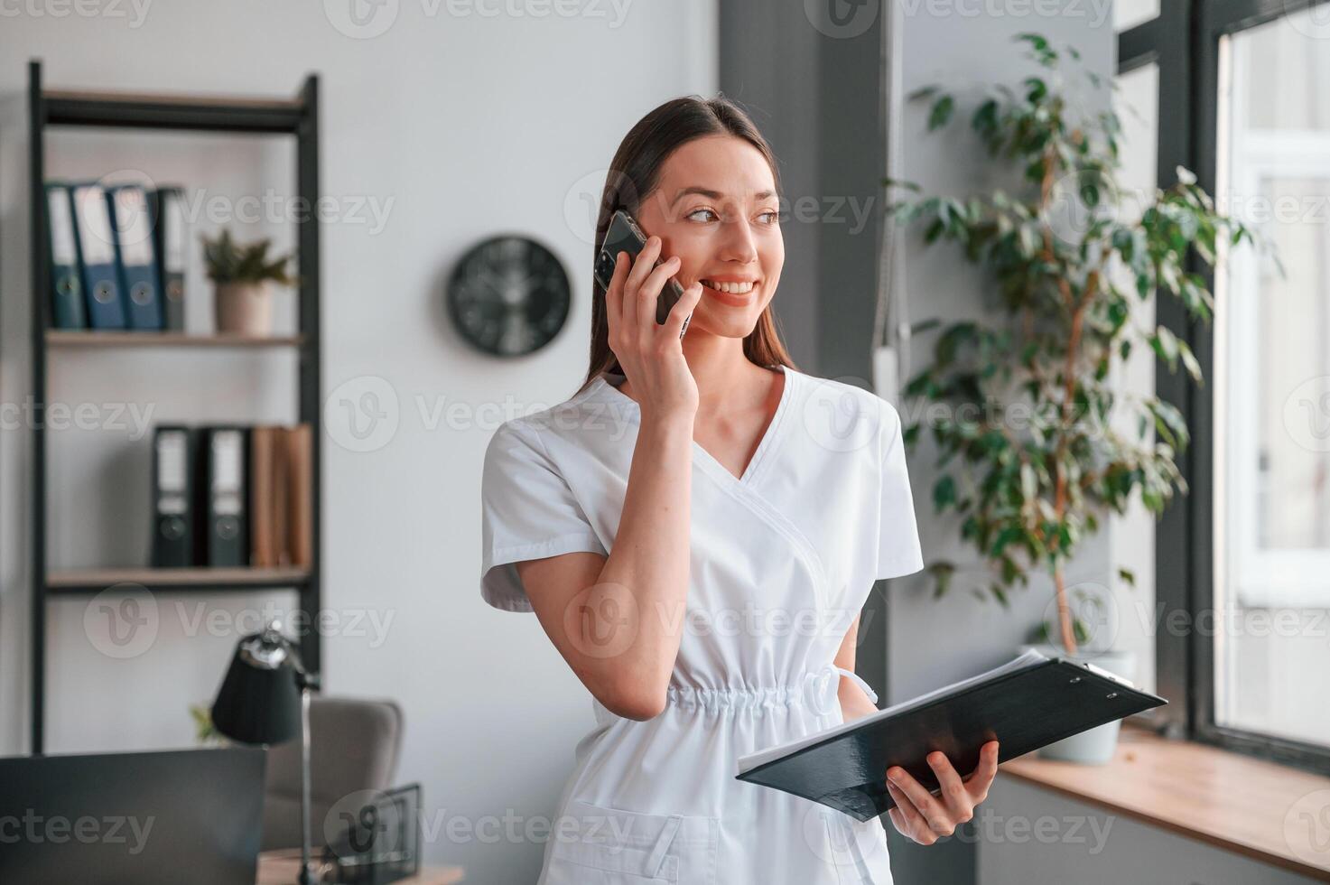 Standing, talking by phone, and holding notepad. Young beautiful woman in white clothes photo
